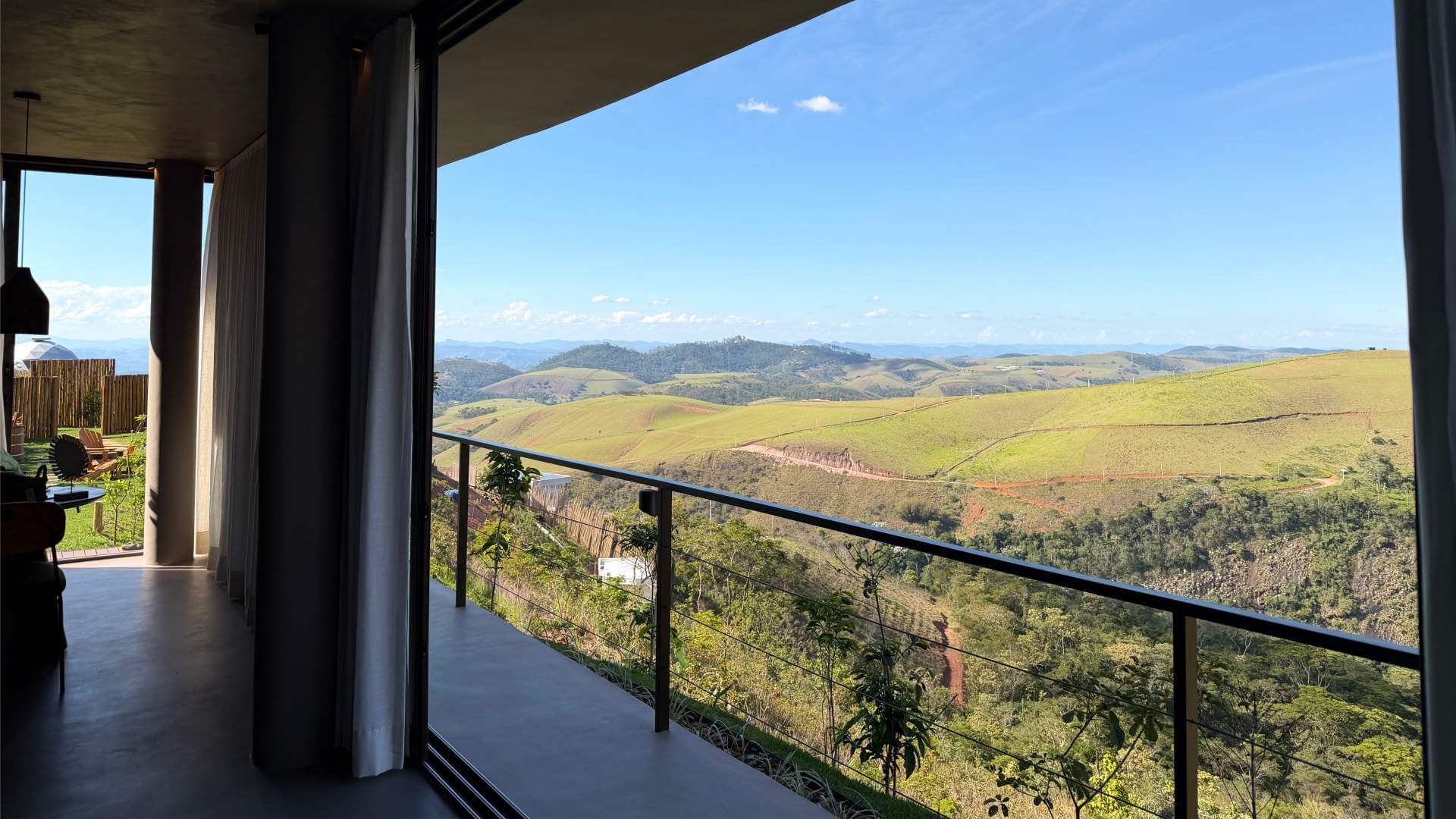 Varanda da Cabana Huna com portas de vidro abertas para o vale verde e as montanhas da Serra Fluminense